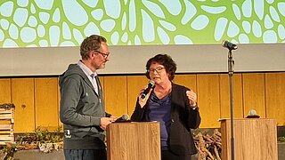 Zwei Personen stehen an Holzpulten auf einer Bühne, eine Person hält ein Mikrofon, im Hintergrund eine grün-weiße abstrakte Musterprojektion