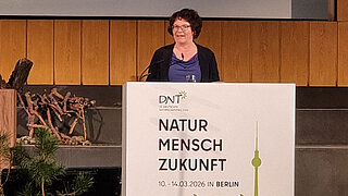Frau steht hinter einem Rednerpult mit dem Schriftzug 'DNT NATUR MENSCH ZUKUNFT 10.-14.03.2026 IN BERLIN' und einer grünen Silhouette von Berliner Wahrzeichen