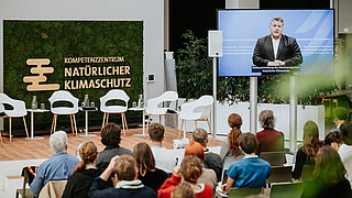 Veranstaltungsbereich mit Publikum und Bildschirm, auf dem Carsten Träger, Parlamentarischer Staatssekretär im Bundesministerium für Umwelt, Klima, Naturschutz und nukleare Sicherheit, zu sehen ist.