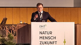 Ein Mann im Anzug steht hinter einem Rednerpult mit dem Schriftzug 'DNT 36. Deutscher Naturschutztag 2026 NATUR MENSCH ZUKUNFT 10.-14.03.2026 IN BERLIN' und einer grünen Silhouette von Berliner Wahrzeichen