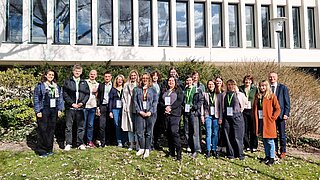 Gruppe von Menschen mit Namensschildern steht auf einer Wiese vor einem Gebäude mit großen Fenstern