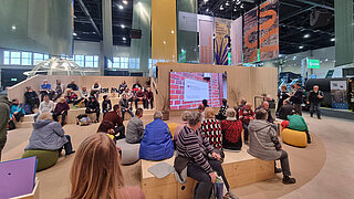 Gruppe von Menschen unterschiedlichen Alters sitzt auf hölzernen Stufen und Sitzkissen in einer großen Halle vor einem Bildschirm mit Präsentation