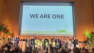 Menschenmenge vor Bühne mit großer Leinwand, auf der 'WE ARE ONE' steht, in einem Saal mit Holzverkleidung und Deckenbeleuchtung