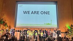 Menschenmenge vor Bühne mit großer Leinwand, auf der 'WE ARE ONE' steht, in einem Saal mit Holzverkleidung und Deckenbeleuchtung