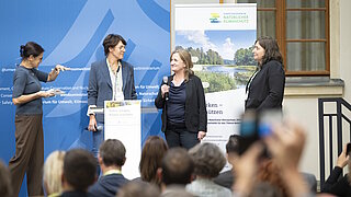 Vier Frauen stehen auf einer Bühne vor Publikum, zwei halten Mikrofone, im Hintergrund Banner mit Text und Landschaftsbild