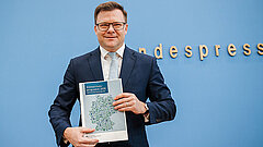 Mann im Anzug hält ein Dokument mit der Aufschrift 'Klimaschutzprogramm 2026' vor einer blauen Wand mit teilweise sichtbarem Schriftzug