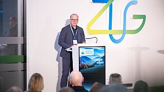 Mann in dunklem Anzug steht am Rednerpult mit Mikrofon vor Publikum, im Hintergrund großes farbiges Logo 'ZUG'.