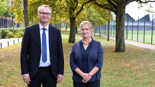 Zwei Personen in Businesskleidung stehen nebeneinander auf einem herbstlichen Parkweg mit Bäumen und einem Zaun im Hintergrund