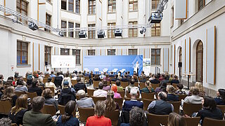 Publikum sitzt in einem hellen Innenhof vor einer Bühne mit blauer Rückwand und mehreren weißen Stühlen