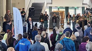 Menschenmenge in einem hellen Innenraum vor einer Treppe mit einem Redner und einem Banner mit Text