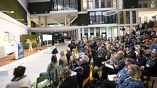 Vortragender steht auf Bühne vor Publikum in modernem, mehrstöckigem Veranstaltungsraum mit großen Fenstern und grünen Wandflächen.