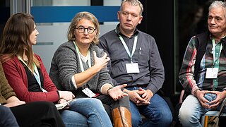 Mehrere Personen sitzen in einem Stuhlkreis, eine Frau mit grauem Pullover und braunen Stiefeln gestikuliert während der Diskussion.