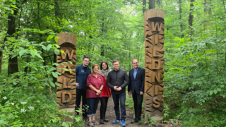 Mehrere Personen (ungter ihnen Carsten Schneider) stehen im Wald. Rechts und links von ihnen befinden sich zwei geschnitzte Holzsäulen mit den Aufschriften "Wald" und "Wildnis". 