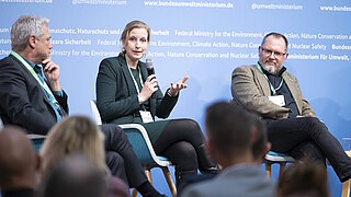 Drei Personen sitzen auf einem Podium vor einem blauen Hintergrund mit Texten zum Bundesumweltministerium, eine Frau spricht in ein Mikrofon, die anderen beiden hören zu
