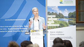 Frau mit kurzem hellgrauem Haar steht an einem Rednerpult und hält ein Smartphone, im Hintergrund Banner mit Texten zum Natürlichen Klimaschutz