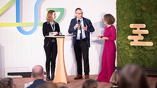 Drei Personen auf einer Bühne beim ANK Fachforum 2025, zwei Frauen und ein Mann, der Mann hält ein Mikrofon, eine Frau trägt ein langes rotes Kleid, im Hintergrund ein grünes Moosbild mit einem stilisierten Logo.