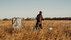 Person steht in einem weiten Feld mit hohem Gras neben einem großen, transparenten Behälter mit blauen Halterungen und hält ein Tablet oder ähnliches Gerät in den Händen