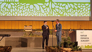 Zwei Männer in Anzügen stehen auf einer Bühne vor einer Leinwand mit grünem Logo und Schrift 'NATUR MENSCH ZUKUNFT 10.-14.03.2026 IN BERLIN' und stilisierter Berliner Skyline
