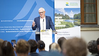 Ein Mann mit Brille und grauem Haar steht an einem Rednerpult und spricht vor einem Publikum, im Hintergrund sind Banner mit Texten zum Klimaschutz zu sehen.