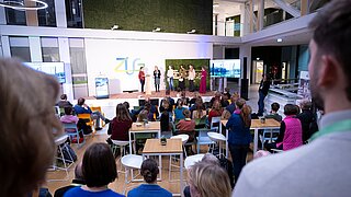 Mehrere Personen stehen auf einer Bühne vor einer Wand mit dem Logo 'ZUG' und sprechen zu einem Publikum in einem modernen, hellen Veranstaltungsraum.