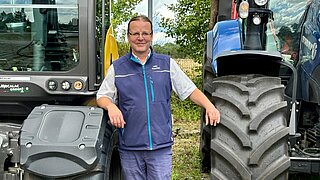 Mann mit Brille und blauem Weste lehnt an großen Reifen von zwei landwirtschaftlichen Fahrzeugen im Freien.
