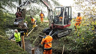Eine Gruppe von Menschen arbeitet an einem Bach mit Bagger 