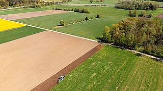 Luftaufnahme einer landwirtschaftlich geprägten Landschaft mit Feldern, Wiesen und Baumgruppen. Im Vordergrund bearbeitet ein Traktor den Rand eines Ackers; im Hintergrund sind weitere Felder und eine Ortschaft zu sehen.