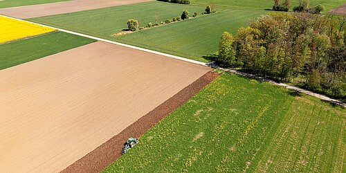 Luftaufnahme einer landwirtschaftlich geprägten Landschaft mit Feldern, Wiesen und Baumgruppen. Im Vordergrund bearbeitet ein Traktor den Rand eines Ackers; im Hintergrund sind weitere Felder und eine Ortschaft zu sehen.