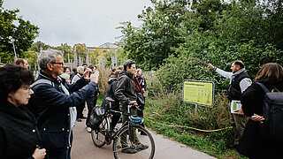 Eine Gruppe von Menschen steht im Park. Eine Person erklärt etwas vor einem Baum.