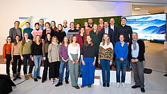 Gruppe von Menschen steht in mehreren Reihen vor einer Bühne mit einem Bildschirm im Hintergrund