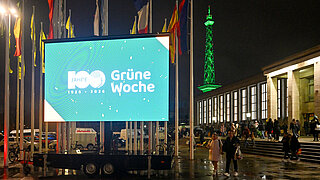Großes beleuchtetes Display mit dem Schriftzug '100 Jahre 1926-2026 Grüne Woche' vor einem Gebäude mit vielen Menschen und einem grünen beleuchteten Turm im Hintergrund