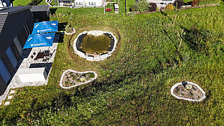 Luftaufnahme eines naturnah gestalteten Firmengeländes im Südschwarzwald mit Teich, zwei kleinen Blumenbeeten und Sitzbereich mit blauen Sonnenschirmen.