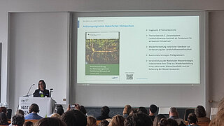 Vortragende Person steht an einem Rednerpult vor einer Leinwand mit einer Präsentationsfolie zum Aktionsprogramm Natürlicher Klimaschutz in einem voll besetzten Seminarraum