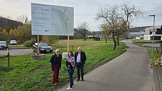 3 Personen stehen auf einem Weg. Im Hintergrund sieht man ein Schild zur Projektplanung