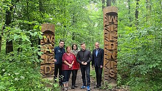 Fünf Personen stehen zwischen zwei hohen Holzsäulen mit geschnitzten Buchstaben in einem grünen Wald.