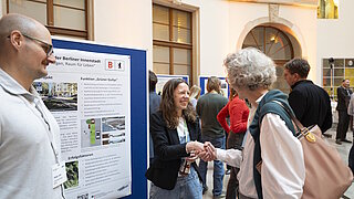 Mehrere Personen unterhalten sich vor einer Ausstellungstafel in einem hellen Innenraum mit hohen Fenstern und Rundbogen