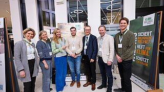 Sieben Personen stehen in einem hellen Raum vor Bannern