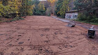 Großflächig ebenerdeter Boden mit zwei Baumaschinen vor bewaldetem Hintergrund und einem Gebäude mit Graffiti