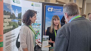 Drei Personen mit Namensschildern stehen an einem Messestand mit Bannern zu Naturschutz und Klimaschutz in einem hellen Raum mit Säulen