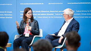 Zwei Personen sitzen auf einem Podium, die linke Person spricht in ein Mikrofon, die rechte hört zu