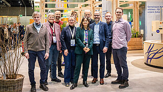 Gruppe von acht Personen steht in einer Halle mit Holzstrukturen und Informationsständen im Hintergrund
