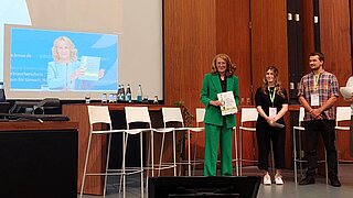 Veranstaltungsteilnehmende auf der Bühne, Steffi Lemke ist per Bildschirm bei der Veranstaltung zugeschaltet