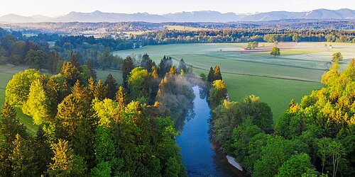 Ein Fluss, der durch einen Wald fließt. Im Hintergrund sind Felder und Berge zu sehen.