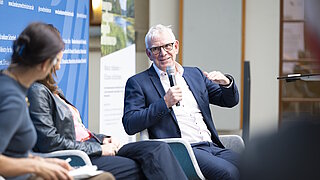 Ein älterer Mann mit Brille und Anzug spricht in ein Mikrofon während einer Podiumsdiskussion, zwei weitere Personen sitzen neben ihm und hören zu