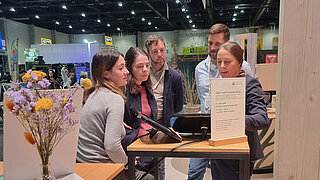 Gruppe von vier Personen an einem Messestand mit einem kleinen Tisch, auf dem ein Schild und ein Tablet stehen, im Hintergrund weitere Messestände und Besucher*innen