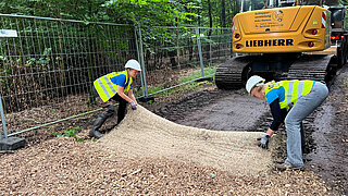 Zwei Personen mit Schutzhelmen und Warnwesten rollen eine Erosionsschutzmatte auf einem mit Holzschnitzeln bedeckten Boden aus, im Hintergrund ein gelber Bagger und ein Bauzaun